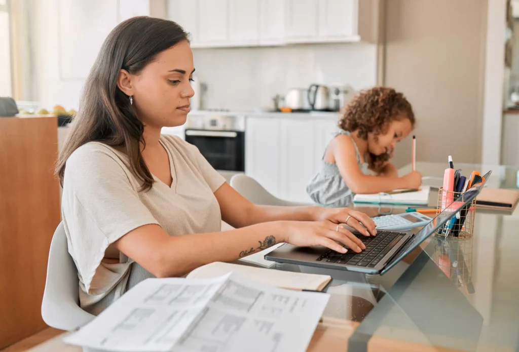 Mother working on a laptop at home while her daughter does homework beside her, representing child care support from Workforce Solutions Capital Area